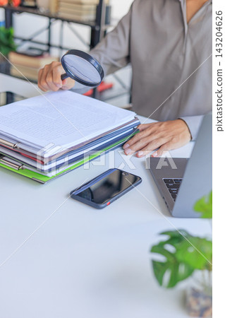 Professional closely inspecting a stack of organized documents and file folders on a desk with a magnifying glass, symbolizing a detailed financial audit, document review, or fraud investigation. Professional closely inspecting a stack of organized documents and file folders on a desk with a magnifying glass, symbolizing a detailed financial audit, document review, or fraud investigation. 143204626
