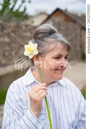 Smiling mature woman with natural gray hair holding a white daffodil beside her face outdoors. Positive aging self confidence natural beauty and midlife lifestyle Smiling mature woman with natural gray hair holding a white daffodil beside her face outdoors. Positive aging self confidence natural beauty and midlife lifestyle 143205002