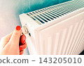 Woman adjusts radiator thermostat for home heating, temperature control, warmth and energy saving during cold winter season. Hand turns valve on white radiator to regulate indoor heat, reduce energy 143205010
