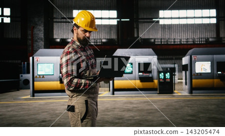 Professional engineer walking through heavy industry workshop in factory, reviewing equipment and metalwork quality on his laptop. Industrial engineering plant features manufacturing production. Professional engineer walking through heavy industry workshop in factory, reviewing equipment and metalwork quality on his laptop. Industrial engineering plant features manufacturing production. 143205474