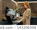 Middle aged businesswoman making morning coffee in office kitchen and checking working plans on laptop . Female preparing hot drink with espresso machine. Corporate lifestyle daily routine concept. 143206308
