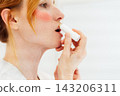 Close up side view Middle aged woman applies lip balm looking in mirror in white bathroom. SPF Lip Balm for lips kin protection. Home beauty treatment and skin care routine. 143206311