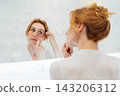 Self Cosmetology, skin care, face treatment, natural beauty concept. Middle aged woman in pajamas looking in mirror and applying pink moisturizing patches on eyes in the bathroom. Home Beauty routine. 143206312