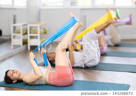 Sports girl with women and men of different ages doing stretch exercises for legs with resistance bands while lying on mats in gym Sports girl with women and men of different ages doing stretch exercises for legs with resistance bands while lying on mats in gym 143206673