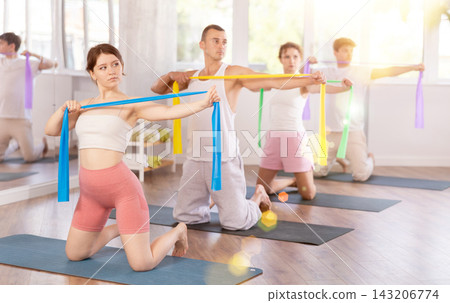 Concentrated sporty young girl exercising with group, kneeling on mat and performing exercise with pilates resistance bands in fitness studio Concentrated sporty young girl exercising with group, kneeling on mat and performing exercise with pilates resistance bands in fitness studio 143206774