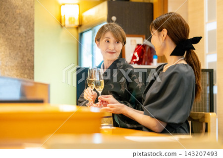 一對母女在壽司店舉杯慶祝:一位六十多歲的女士穿著和服,一位三十多歲的女士穿著連衣裙。 一對母女在壽司店舉杯慶祝:一位六十多歲的女士穿著和服,一位三十多歲的女士穿著連衣裙。 143207493