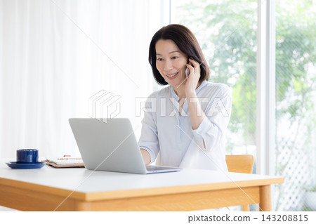 一位中年婦女遠距辦公,一邊看著電腦一邊打電話。 一位中年婦女遠距辦公,一邊看著電腦一邊打電話。 143208815