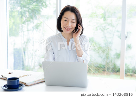 一位中年婦女遠距辦公，一邊看著電腦一邊打電話。 143208816