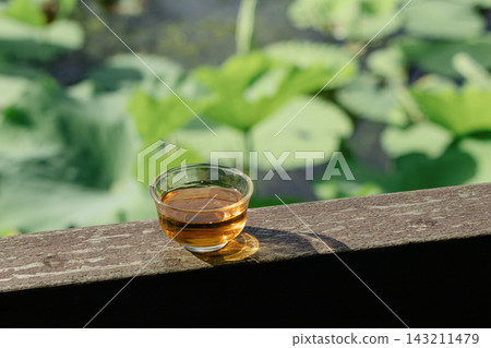 Small glass cup of amber tea sitting on a rustic wooden railing with a blurred background of green lotus leaves in a garden pond Small glass cup of amber tea sitting on a rustic wooden railing with a blurred background of green lotus leaves in a garden pond 143211479