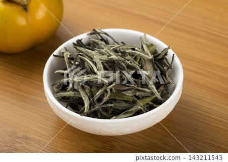 Dry white tea leaves in a white ceramic bowl on a wooden table, silver needle tea buds closeup 143211543