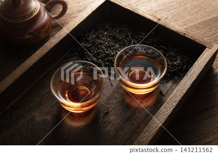 Traditional Chinese tea ceremony featuring two small glass cups of dark oolong tea, loose tea leaves, and a clay teapot on a Traditional Chinese tea ceremony featuring two small glass cups of dark oolong tea, loose tea leaves, and a clay teapot on a 143211562