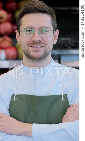 Vertical video. Close up portrait of happy man worker in glasses wearing apron standing in supermarket and looking at camera. Retail business. Vertical video. Close up portrait of happy man worker in glasses wearing apron standing in supermarket and looking at camera. Retail business. 143214109