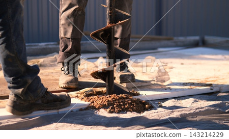 Boots and protective tarp frame close view of workers drilling soil with helical auger, scattering loose earth on house construction site during daylight building progress Boots and protective tarp frame close view of workers drilling soil with helical auger, scattering loose earth on house construction site during daylight building progress 143214229