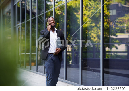 Businessman walking outside office building holding laptop talking on phone Businessman walking outside office building holding laptop talking on phone 143222201