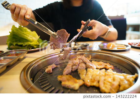 Asian woman using tongs and scissors cutting grilled pork at Korean BBQ restaurant Asian woman using tongs and scissors cutting grilled pork at Korean BBQ restaurant 143222224