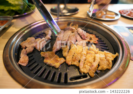 Asian woman using tongs and scissors cutting grilled pork at Korean BBQ restaurant Asian woman using tongs and scissors cutting grilled pork at Korean BBQ restaurant 143222226
