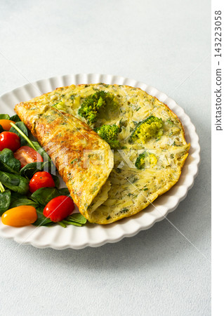 A potato omelette made with eggs and mixed greens is served on a white plate. Spinach, cherry tomatoes, and broccoli are arranged on the side for a healthy breakfast option A potato omelette made with eggs and mixed greens is served on a white plate. Spinach, cherry tomatoes, and broccoli are arranged on the side for a healthy breakfast option 143223058