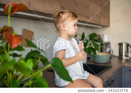 Happy baby girl sitting at the table in the kitchen and drinking milk Happy baby girl sitting at the table in the kitchen and drinking milk 143223778