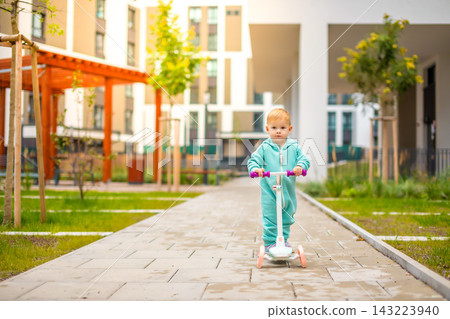 Cute little toddler girl in blue overalls riding on kick scooter. Happy healthy lovely baby child having fun in the city. Active kid on cold day outdoors. Cute little toddler girl in blue overalls riding on kick scooter. Happy healthy lovely baby child having fun in the city. Active kid on cold day outdoors. 143223940