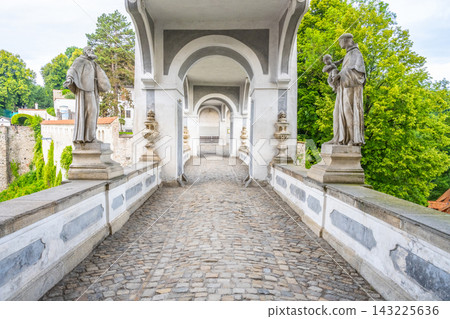 The Coat Bridge connects the Upper Castle and the Baroque Castle Theatre in Cesky Krumlov. Statues line the sides of the stone pathway surrounded by green trees. The Coat Bridge connects the Upper Castle and the Baroque Castle Theatre in Cesky Krumlov. Statues line the sides of the stone pathway surrounded by green trees. 143225636
