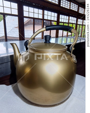 房間裡擺放著一個昭和時代的復古風格的大型金色鋁製水壺,放在托盤上,房間裡還有木框窗戶。 房間裡擺放著一個昭和時代的復古風格的大型金色鋁製水壺,放在托盤上,房間裡還有木框窗戶。 143226144