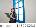 Young professional woman standing beside bright blue window frame holding tablet in modern office interior with soft daylight, smiling confidently, wearing tailored business suit and white blouse 143226238