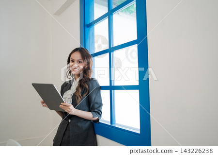 Young professional woman standing beside bright blue window frame holding tablet in modern office interior with soft daylight, smiling confidently, wearing tailored business suit and white blouse Young professional woman standing beside bright blue window frame holding tablet in modern office interior with soft daylight, smiling confidently, wearing tailored business suit and white blouse 143226238