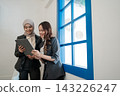 Two professional women sharing tablet beside bright blue window in modern office, smiling during collaborative work, planning, review, and friendly business discussion indoors 143226247