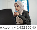 Thoughtful young woman in hijab working at laptop with focused expression in bright office interior, hands clasped near face, modern professional portrait with soft natural light 143226249