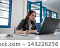 Focused young businesswoman writing beside laptop in bright modern office, working through documents with concentrated expression, blue framed windows, clean workspace, professional daytime setting 143226256