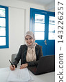 Smiling woman in hijab writing notes at desk beside laptop in bright office with blue windows, professional portrait showing focused work, education, planning, and modern business environment 143226257