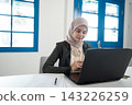 Focused young woman in hijab working on laptop at bright modern office desk with blue windows, papers, notebook, and soft daylight, showing professional online business productivity 143226259