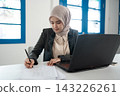 Young Muslim businesswoman in hijab writing on paper beside laptop at bright office desk, focused on paperwork, planning, and administrative work in modern workspace 143226261