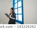 Smiling businesswoman using digital tablet beside bright blue window in modern office interior with clean white wall, elegant suit, soft daylight, professional workspace, calm corporate atmosphere 143226262