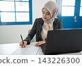 Muslim businesswoman in hijab writing notes at office desk beside laptop under bright window light, focused on paperwork, planning, administration, and professional tasks 143226300