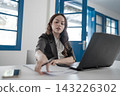 Young businesswoman in modern office reaching across desk beside open laptop and papers, focused on work in bright interior with blue doors and windows 143226302