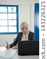 Young woman in hijab writing on document beside laptop in bright office, focused on paperwork and computer work at desk with blue window frames, professional indoor business setting 143226325