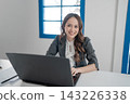 Smiling businesswoman working at laptop in bright office with window light, wearing blazer and white blouse, seated at desk with documents, showing confident professional attitude 143226338