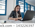 Focused young businesswoman writing notes beside laptop in bright modern office with blue windows, concentrating on paperwork, planning daily tasks, and digital work 143226397