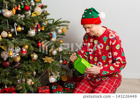 A 53 year old man sits near a decorated Christmas tree on Christmas Day. He is opening a gift while wearing a festive outfit and a holiday hat. Family and joy fill the space around him A 53 year old man sits near a decorated Christmas tree on Christmas Day. He is opening a gift while wearing a festive outfit and a holiday hat. Family and joy fill the space around him 143226449