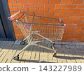 Empty metal shopping cart parked on paved ground against a red brick and grey metal wall. An abandoned cart outside a supermarket 143227989