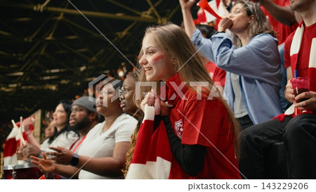 Tense young female football supporter holding red scarf to mouth while watching crucial live match from the stands, women in football. Anxious girl with face paint living intense football match. Tense young female football supporter holding red scarf to mouth while watching crucial live match from the stands, women in football. Anxious girl with face paint living intense football match. 143229206