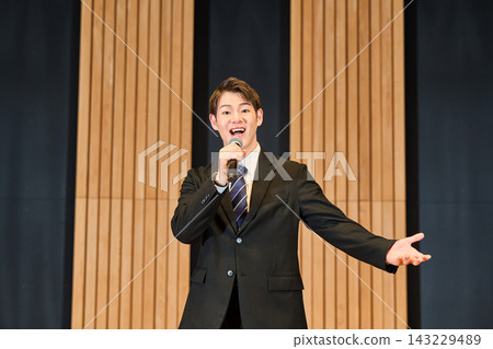 A young man in a suit, holding a microphone in a hall, delivers a presentation or sings a song. A young man in a suit, holding a microphone in a hall, delivers a presentation or sings a song. 143229489