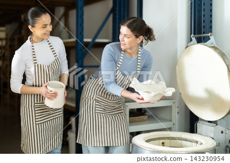 Two female potters take finished ceramic dishes, various mugs and a vase out of oven Two female potters take finished ceramic dishes, various mugs and a vase out of oven 143230954