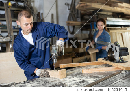 Male employee of carpentry workshop uses drill to make holes in wooden billet 143231045