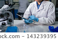 Scientist wearing blue gloves reviews data on tablet beside microscope in modern research laboratory 143239395