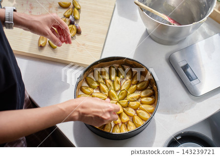 Woman arranging sliced plums on dough while making pie in kitchen. Woman arranging sliced plums on dough while making pie in kitchen. 143239721