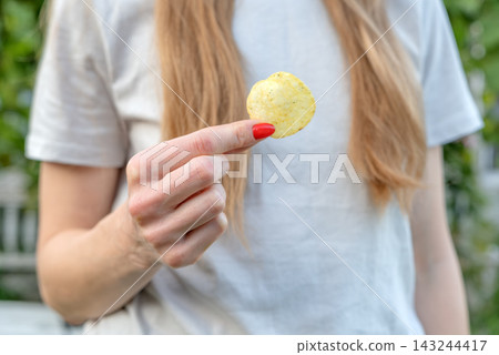 Woman's hand with red nail polish holding up a single golden potato chip, suggesting an indulgent snack or craving for crunchy fast food in a casual outdoor setting Woman's hand with red nail polish holding up a single golden potato chip, suggesting an indulgent snack or craving for crunchy fast food in a casual outdoor setting 143244417