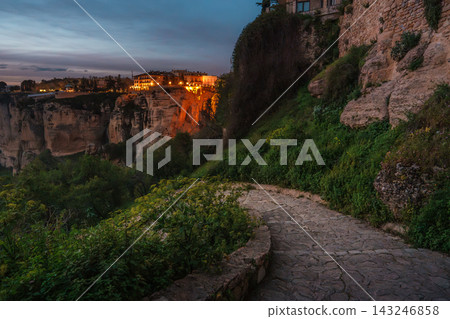 Stone footpath below Ronda cliffs toward illuminated clifftop buildings at dusk Stone footpath below Ronda cliffs toward illuminated clifftop buildings at dusk 143246858