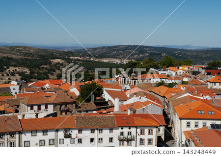 Aerial cityscape of orange tiled rooftops and church bell towers in the old town of Guarda, Portugal, with green hills and wind turbines. Travel and tourism destination in historic Europe. 143248449